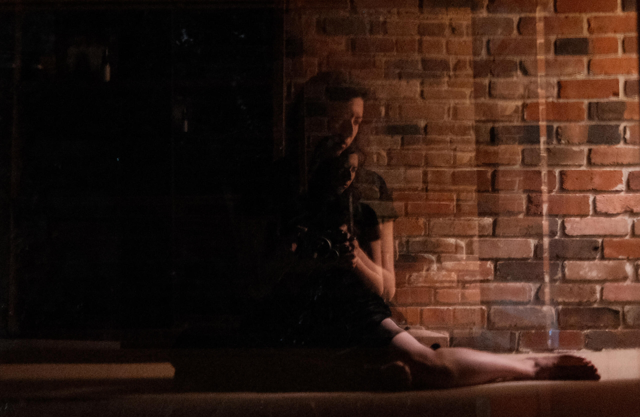 A self portrait photograph of Leanne through a reflective surface that shows multiple repeating reflections, half in shadow, against a brick wall.